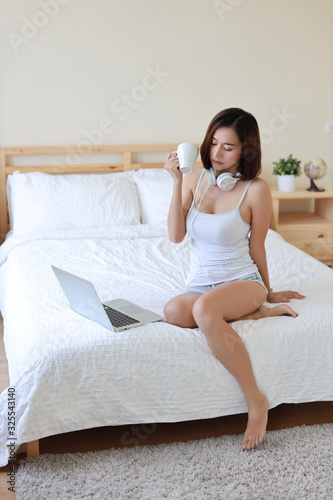 Beautiful and young asian woman in white casual dress with attractive look, lies on bed with computer and headphone. Cut girl and short hair using computer, drinking coffee with happiness in bedroom