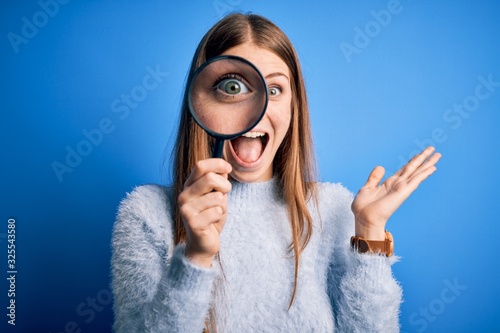 Young beautiful redhead detective woman using magnifying glass over isolated blue background very happy and excited, winner expression celebrating victory screaming with big smile and raised hands photo