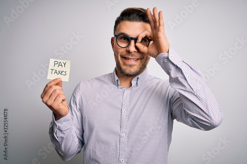 Young business man with blue eyes holding pay taxes word on paper note with happy face smiling doing ok sign with hand on eye looking through fingers © Krakenimages.com