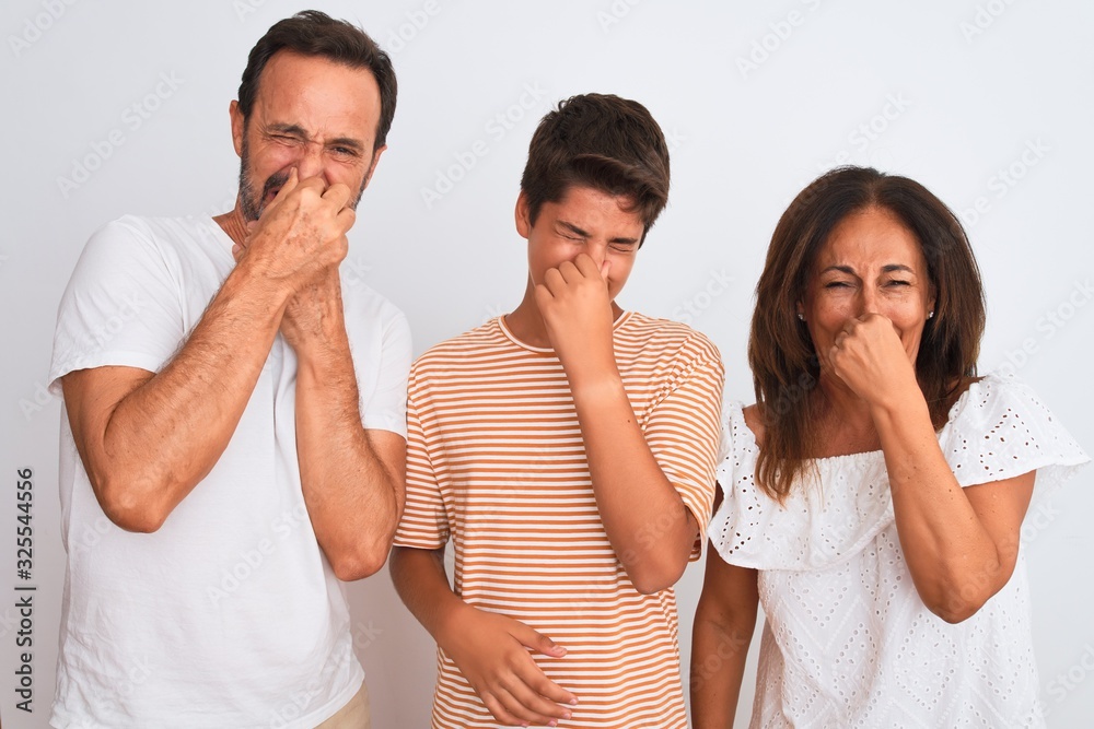 Family of three, mother, father and son standing over white isolated background smelling something stinky and disgusting, intolerable smell, holding breath with fingers on nose. Bad smells concept. Family of three, mother, father and son standing over white isolated background smelling something stinky and disgusting, intolerable smell, holding breath with fingers on nose. Bad smells concept.
