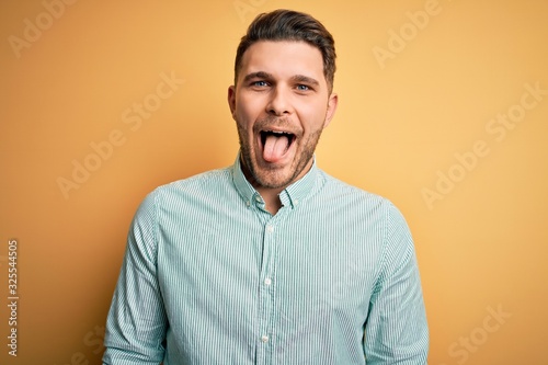 Young business man with blue eyes wearing elegant green shirt over yellow background sticking tongue out happy with funny expression. Emotion concept.