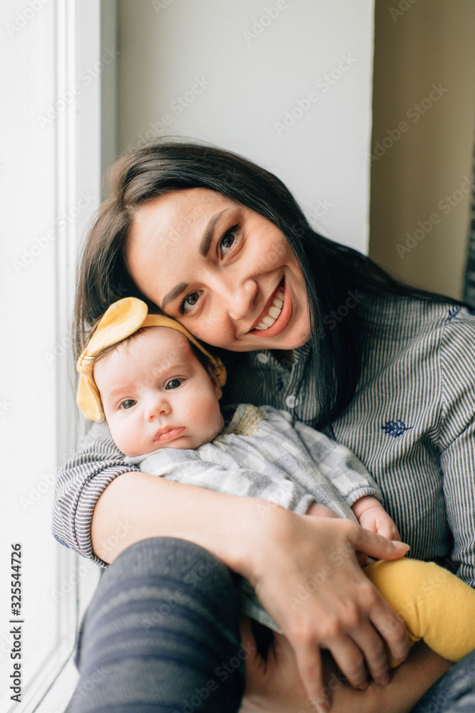 Portrait of beautiful woman with black hair sits on a windowsill with her pretty little child and smiles Portrait of beautiful woman with black hair sits on a windowsill with her pretty little child and smiles