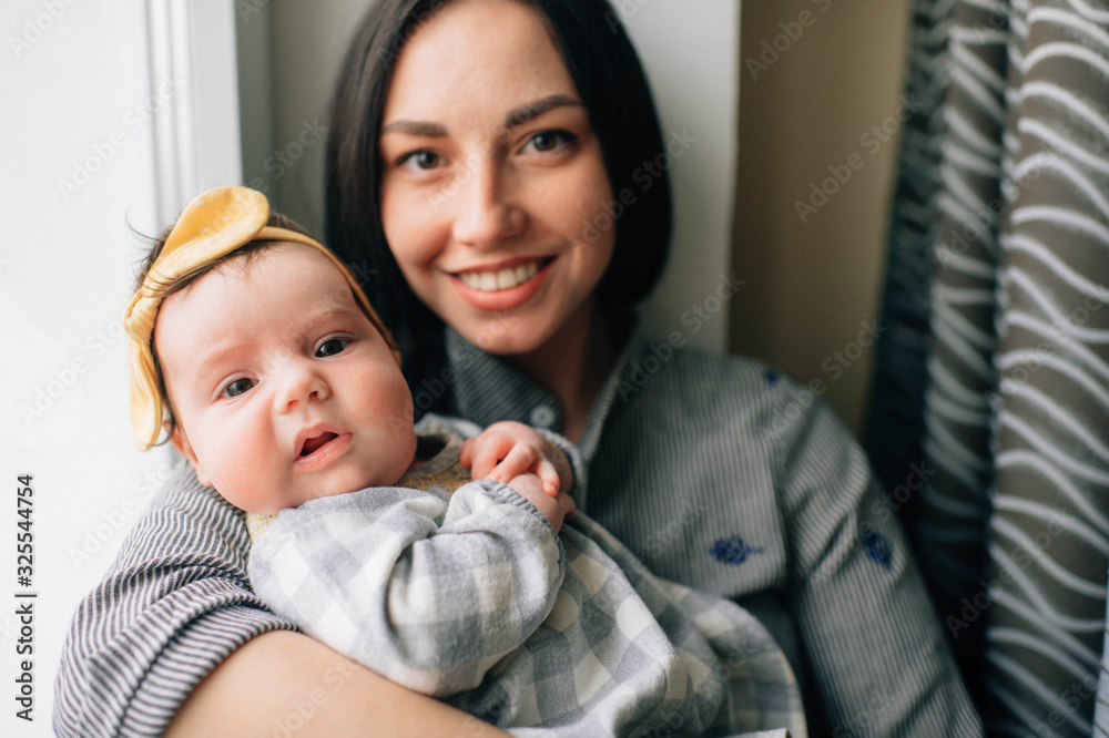 Fototapeta premium Portrait of pretty woman with black hair sits on a windowsill with her pretty little child and smiles