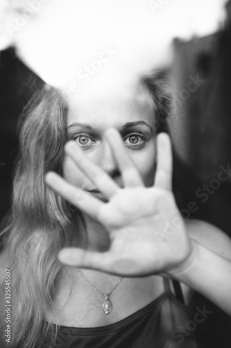 Fototapeta Naklejka Na Ścianę i Meble -  Black and white picture of handsome young girl with fair hair behind glass and covers her face with the hand vand say to stop