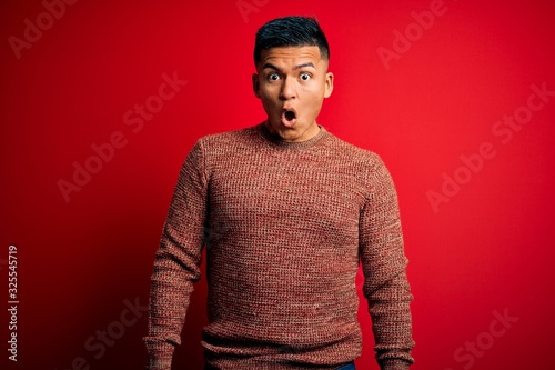 Young handsome latin man wearing casual sweater standing over red background afraid and shocked with surprise expression, fear and excited face.