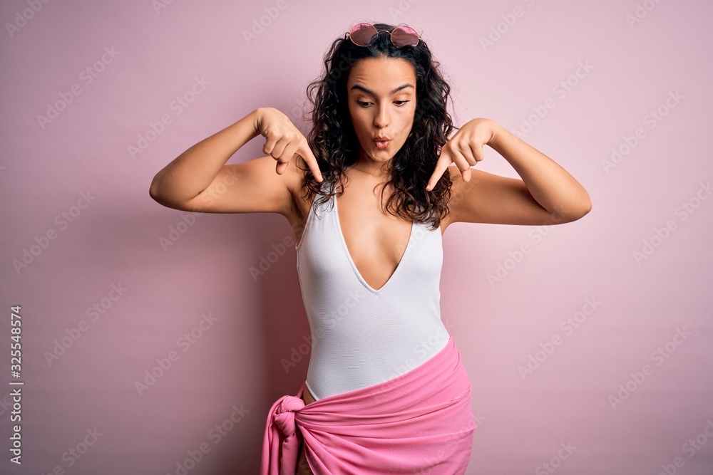 Beautiful woman with curly hair on vacation wearing white swimsuit over pink background Pointing down with fingers showing advertisement, surprised face and open mouth Beautiful woman with curly hair on vacation wearing white swimsuit over pink background Pointing down with fingers showing advertisement, surprised face and open mouth