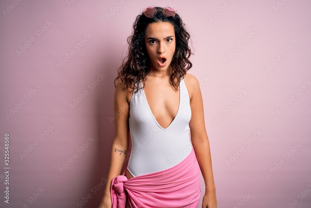 Beautiful woman with curly hair on vacation wearing white swimsuit over pink background In shock face, looking skeptical and sarcastic, surprised with open mouth Beautiful woman with curly hair on vacation wearing white swimsuit over pink background In shock face, looking skeptical and sarcastic, surprised with open mouth