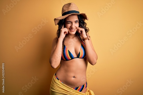 Young beautiful woman with curly hair on vacation wearing bikini and summer hat covering ears with fingers with annoyed expression for the noise of loud music. Deaf concept.