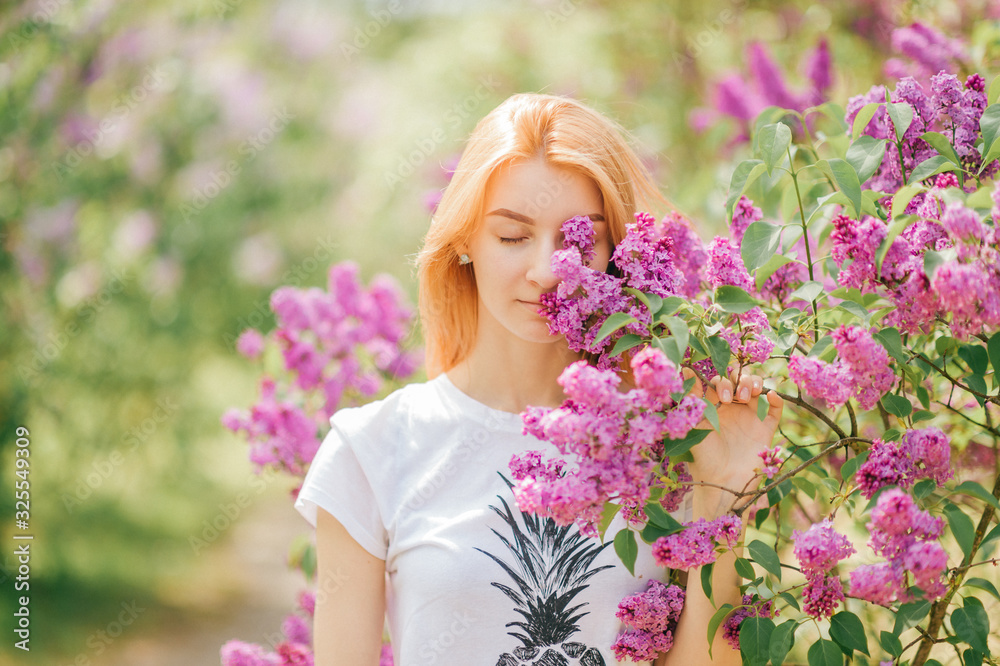 Pretty young female with red hair and fair skin smells lilac in the garden with closed eyes