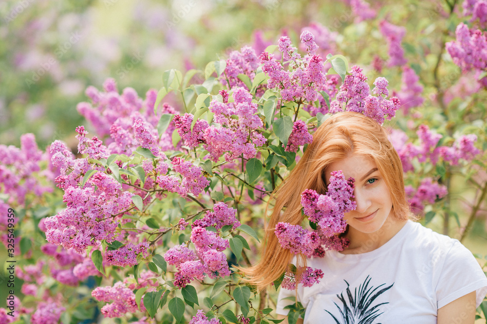 Handsome woman with red hair and pretty smile posing in lilac garden with flowers and enjoys the smell Handsome woman with red hair and pretty smile posing in lilac garden with flowers and enjoys the smell