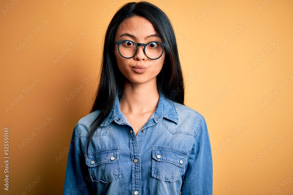 Young beautiful chinese woman wearing casual denim shirt over isolated yellow background making fish face with lips, crazy and comical gesture. Funny expression.