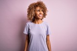 Young beautiful african american woman wearing casual t-shirt standing over pink background looking away to side with smile on face, natural expression. Laughing confident.