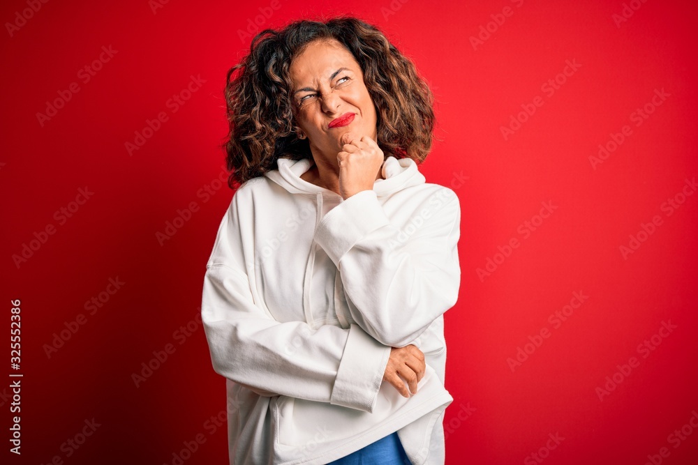 Middle age beautiful sporty woman wearing white sweatshirt over isolated red background with hand on chin thinking about question, pensive expression. Smiling with thoughtful face. Doubt concept.