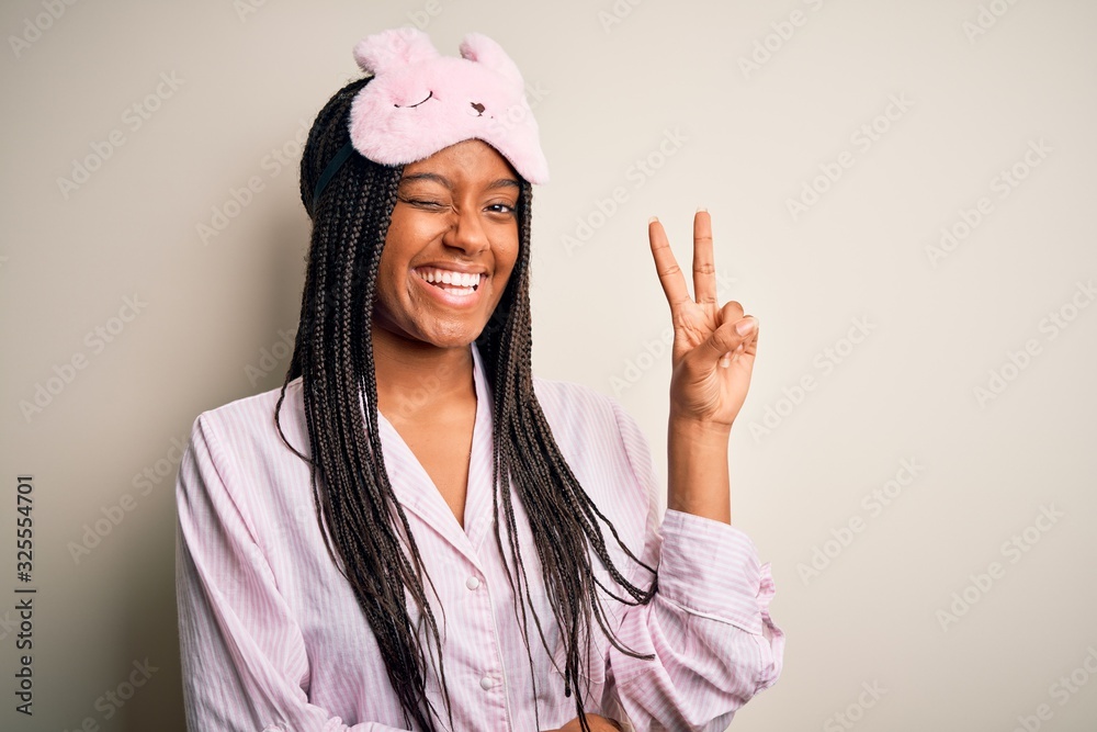 Young african american woman wearing pink pajama and sleep mask over isolated background smiling with happy face winking at the camera doing victory sign. Number two. Young african american woman wearing pink pajama and sleep mask over isolated background smiling with happy face winking at the camera doing victory sign. Number two.