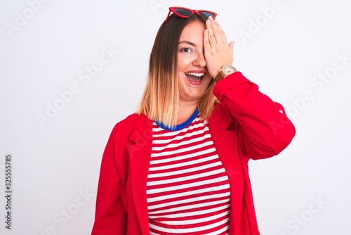 Young beautiful woman wearing striped t-shirt and jacket over isolated white background covering one eye with hand, confident smile on face and surprise emotion.