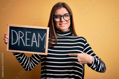Young beautiful woman with blue eyes holding blackboard with dream message word with surprise face pointing finger to himself