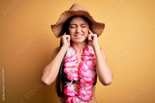 Young beautiful woman with blue eyes on vacation wearing bikini and hawaiian lei covering ears with fingers with annoyed expression for the noise of loud music. Deaf concept.