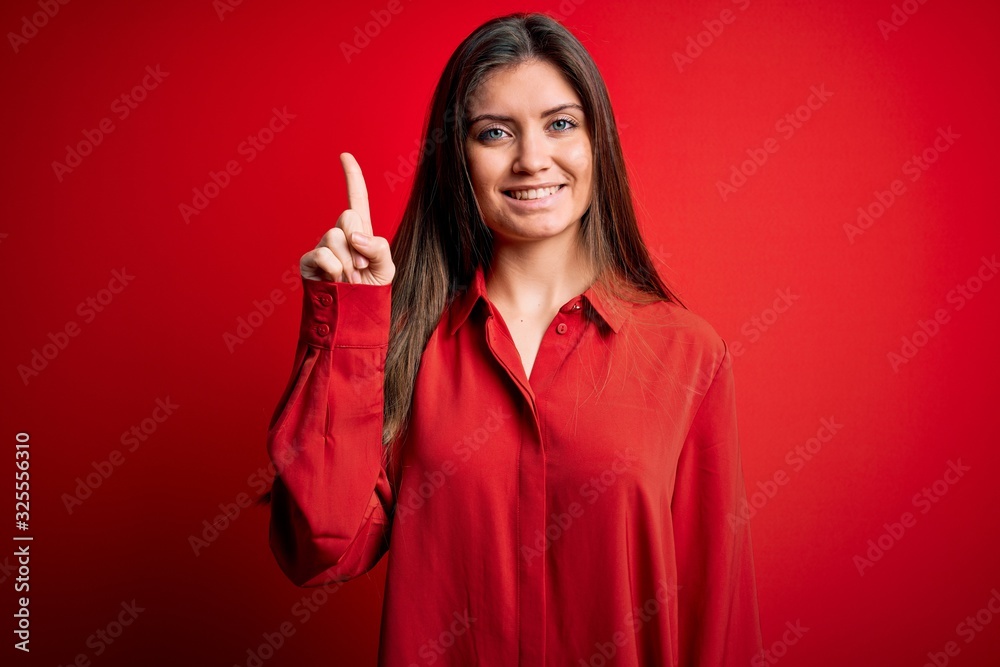 Young beautiful woman with blue eyes wearing casual shirt standing over red background showing and pointing up with finger number one while smiling confident and happy. Young beautiful woman with blue eyes wearing casual shirt standing over red background showing and pointing up with finger number one while smiling confident and happy.