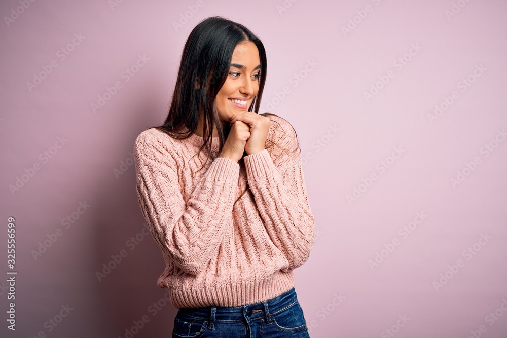 Young beautiful brunette woman wearing casual sweater over isolated pink background laughing nervous and excited with hands on chin looking to the side