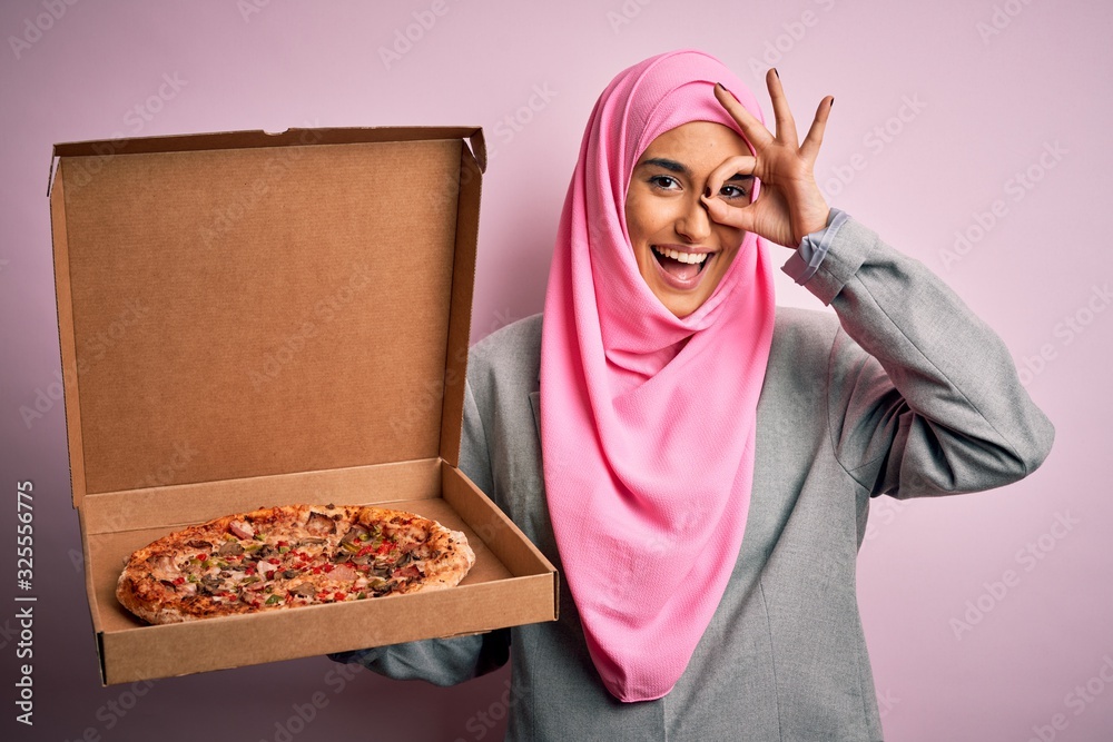 Young beautiful arab woman wearing pink muslim hijab holding delivery box with Italian pizza with happy face smiling doing ok sign with hand on eye looking through fingers Young beautiful arab woman wearing pink muslim hijab holding delivery box with Italian pizza with happy face smiling doing ok sign with hand on eye looking through fingers