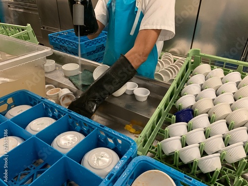 Kitchen porter is wearing long black gloves he is cleaning plates in sink in a commercial galley on the cruise ship.