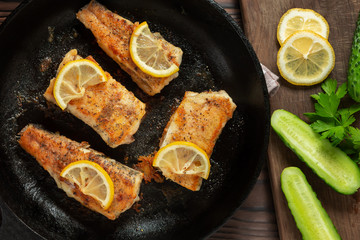 Portioned pieces of fried fish in a black cast-iron pan with fresh cucumbers and herbs. Traditional dinner of a village fisherman, top view, flat lay