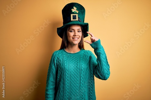 Young beautiful brunette woman wearing green hat with clover celebrating saint patricks day smiling and confident gesturing with hand doing small size sign with fingers looking and the camera. Measure