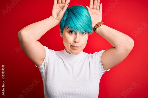 Young beautiful woman with blue fashion hair wearing casual t-shirt over red background Doing bunny ears gesture with hands palms looking cynical and skeptical. Easter rabbit concept. photo