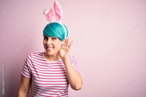 Young woman with fashion blue hair wearing easter rabbit ears over pink background smiling with hand over ear listening an hearing to rumor or gossip. Deafness concept.