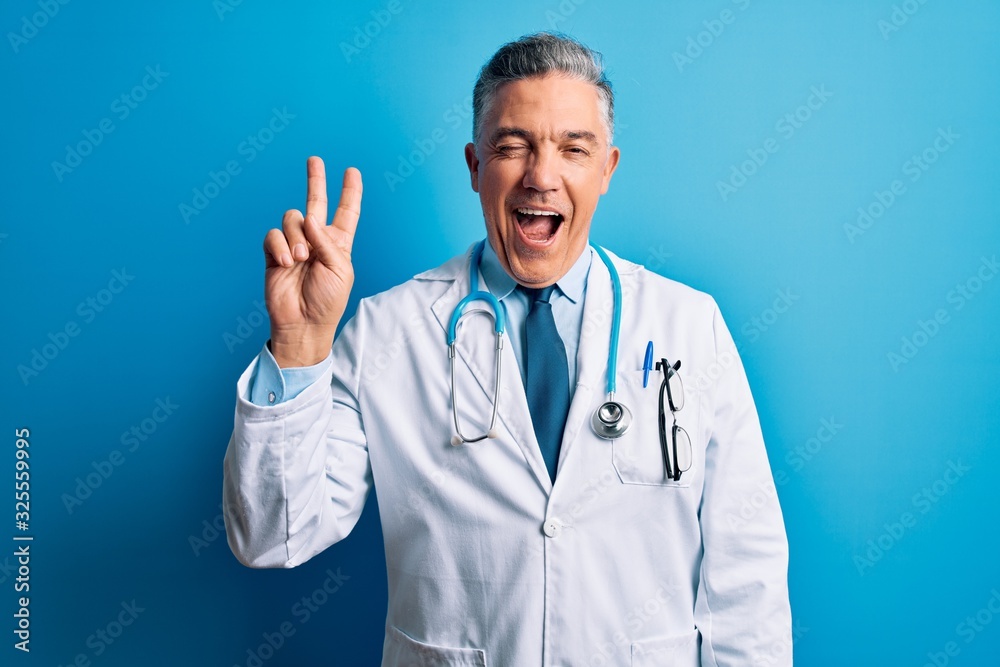 Middle age handsome grey-haired doctor man wearing coat and blue stethoscope smiling with happy face winking at the camera doing victory sign with fingers. Number two. Middle age handsome grey-haired doctor man wearing coat and blue stethoscope smiling with happy face winking at the camera doing victory sign with fingers. Number two.