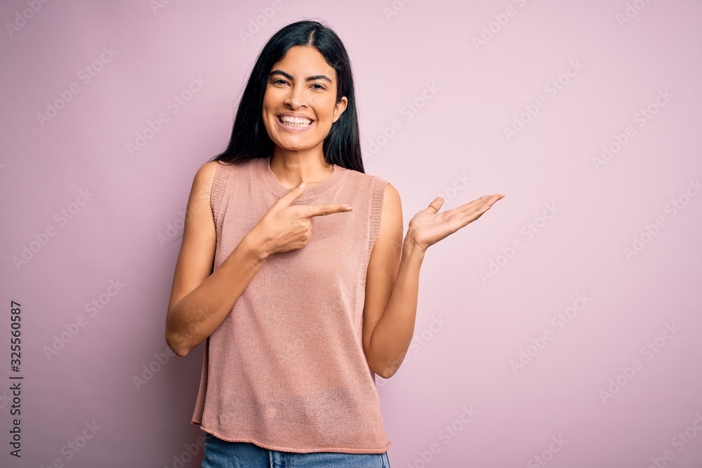 Young beautiful hispanic fashion woman wearing casual sweater over pink background amazed and smiling to the camera while presenting with hand and pointing with finger. Young beautiful hispanic fashion woman wearing casual sweater over pink background amazed and smiling to the camera while presenting with hand and pointing with finger.