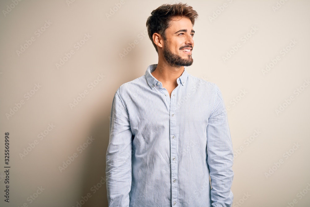 Young handsome man with beard wearing striped shirt standing over white background looking away to side with smile on face, natural expression. Laughing confident. Young handsome man with beard wearing striped shirt standing over white background looking away to side with smile on face, natural expression. Laughing confident.