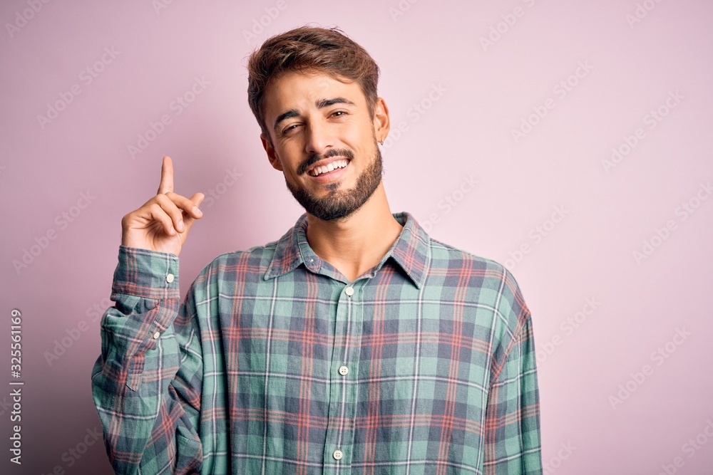 Naklejka premium Young handsome man with beard wearing casual shirt standing over pink background with a big smile on face, pointing with hand and finger to the side looking at the camera.