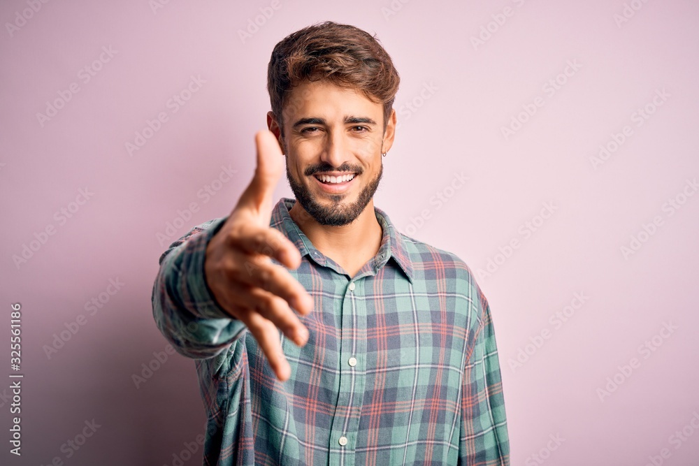 Young handsome man with beard wearing casual shirt standing over pink background smiling friendly offering handshake as greeting and welcoming. Successful business. Young handsome man with beard wearing casual shirt standing over pink background smiling friendly offering handshake as greeting and welcoming. Successful business.