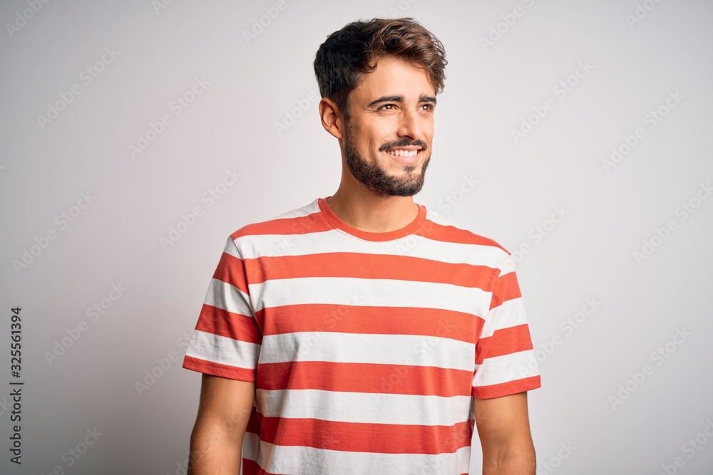 Young handsome man with beard wearing striped t-shirt standing over white background looking away to side with smile on face, natural expression. Laughing confident. Young handsome man with beard wearing striped t-shirt standing over white background looking away to side with smile on face, natural expression. Laughing confident.