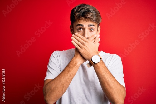 Young handsome man with beard wearing casual t-shirt standing over red background shocked covering mouth with hands for mistake. Secret concept.
