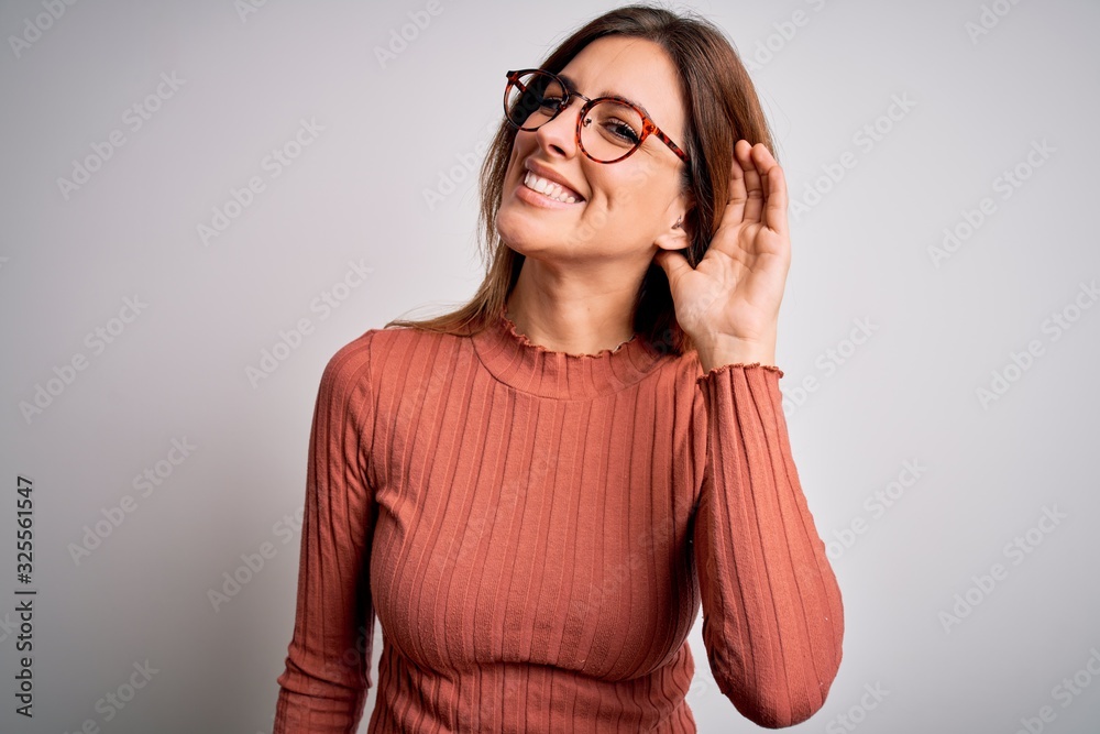 Young beautiful brunette woman wearing casual sweater and glasses over white background smiling with hand over ear listening an hearing to rumor or gossip. Deafness concept. Young beautiful brunette woman wearing casual sweater and glasses over white background smiling with hand over ear listening an hearing to rumor or gossip. Deafness concept.