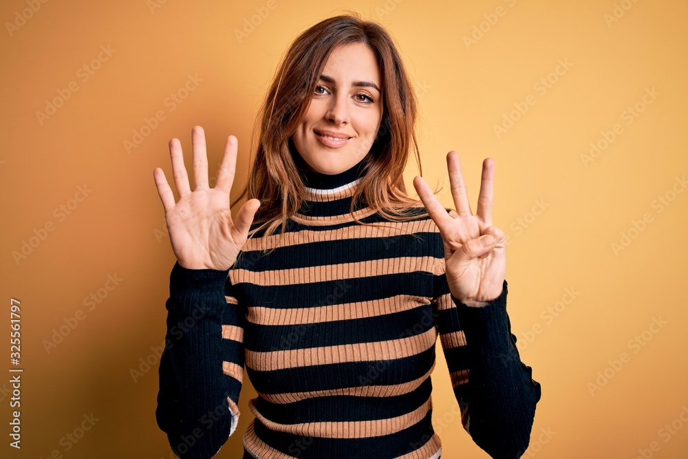 Young beautiful brunette woman wearing striped turtleneck sweater over yellow background showing and pointing up with fingers number eight while smiling confident and happy. Young beautiful brunette woman wearing striped turtleneck sweater over yellow background showing and pointing up with fingers number eight while smiling confident and happy.
