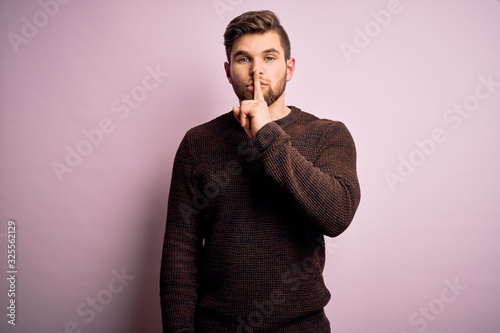 Young blond man with beard and blue eyes wearing casual sweater over pink background asking to be quiet with finger on lips. Silence and secret concept. © Krakenimages.com
