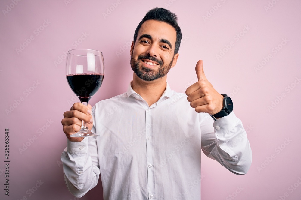 Young handsome man with beard drinking glass of red wine over isolated pink background happy with big smile doing ok sign, thumb up with fingers, excellent sign Young handsome man with beard drinking glass of red wine over isolated pink background happy with big smile doing ok sign, thumb up with fingers, excellent sign