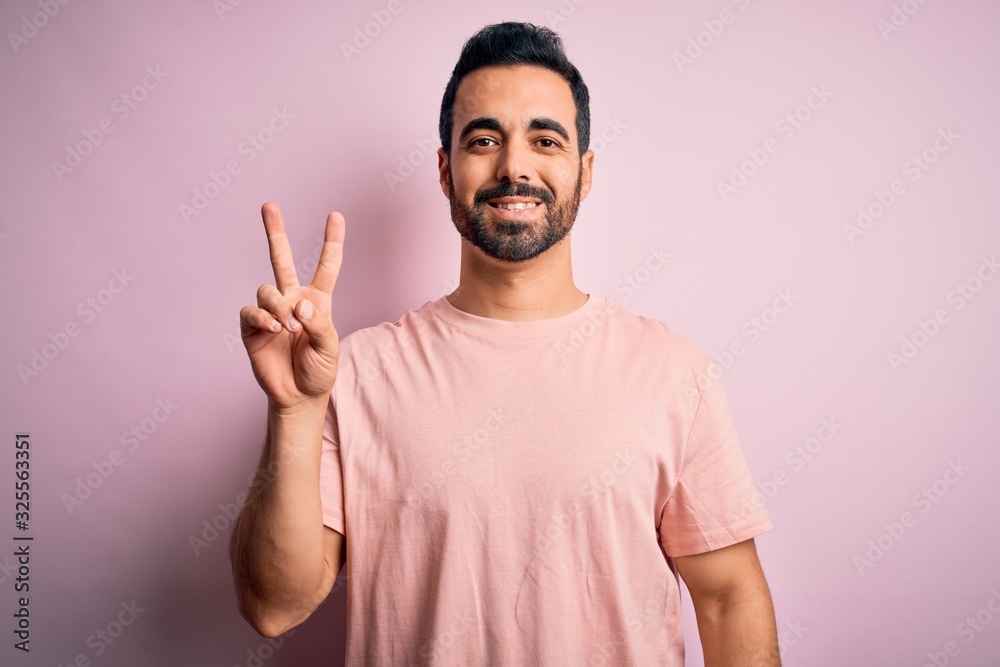 Young handsome man with beard wearing casual t-shirt standing over pink background smiling with happy face winking at the camera doing victory sign. Number two. Young handsome man with beard wearing casual t-shirt standing over pink background smiling with happy face winking at the camera doing victory sign. Number two.