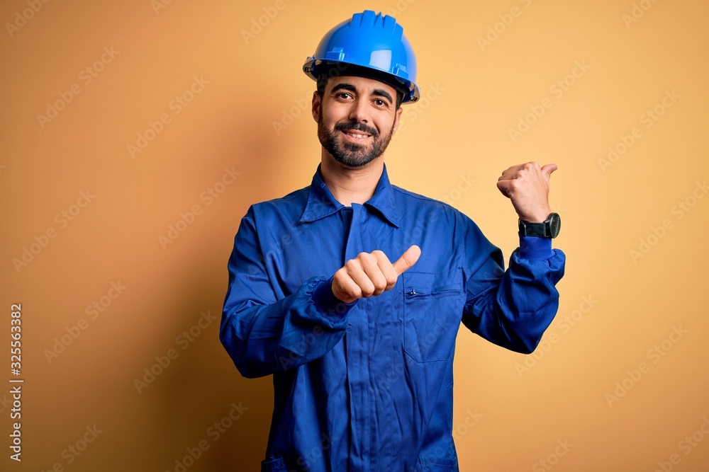 Mechanic man with beard wearing blue uniform and safety helmet over yellow background Pointing to the back behind with hand and thumbs up, smiling confident