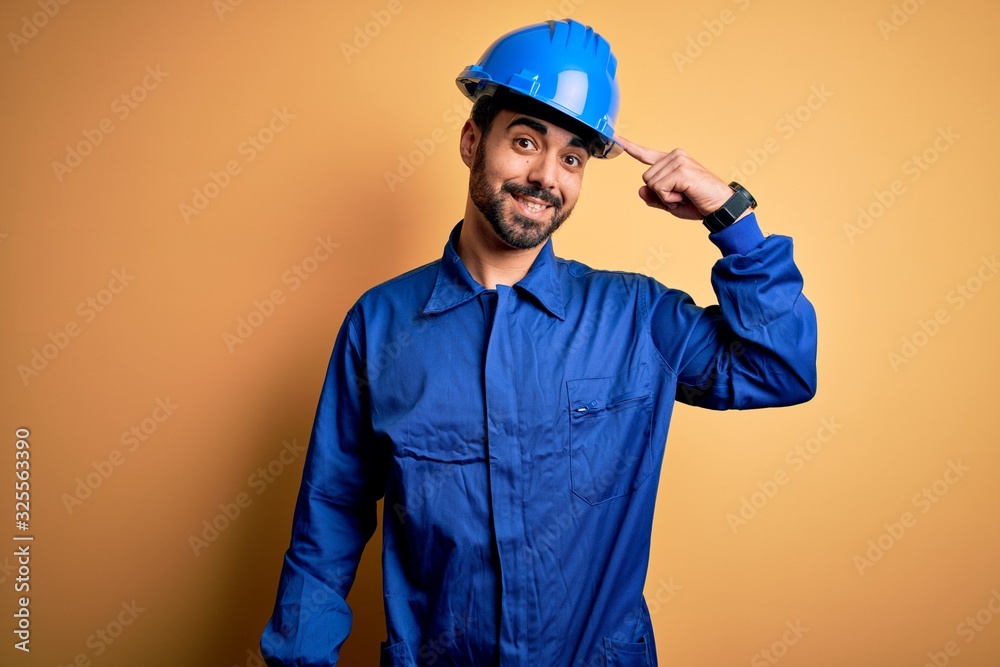 Mechanic man with beard wearing blue uniform and safety helmet over yellow background Smiling pointing to head with one finger, great idea or thought, good memory