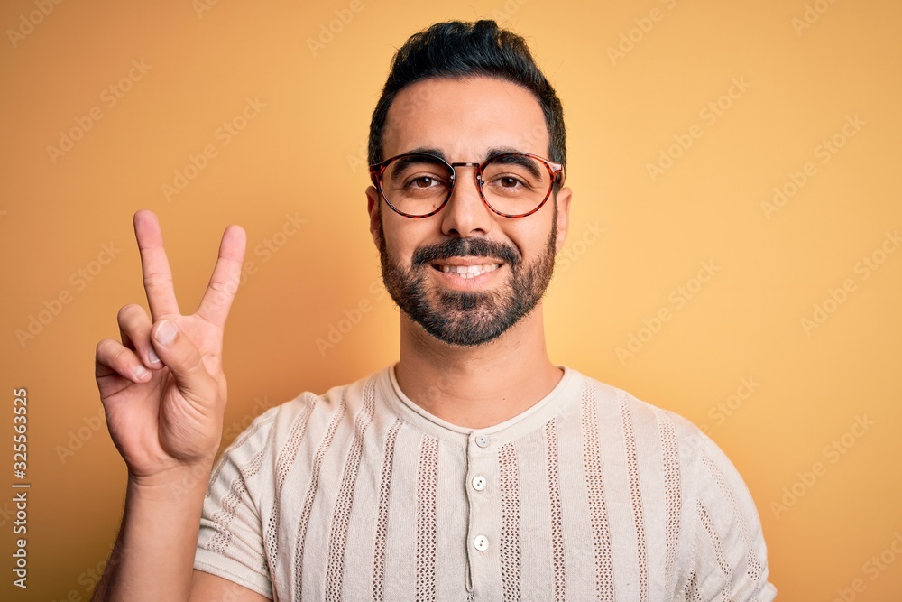 Young handsome man with beard wearing casual t-shirt and glasses over yellow background smiling with happy face winking at the camera doing victory sign. Number two. Young handsome man with beard wearing casual t-shirt and glasses over yellow background smiling with happy face winking at the camera doing victory sign. Number two.