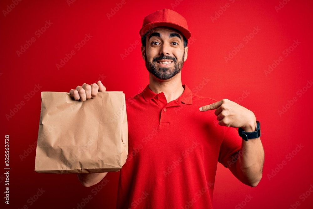 Young handsome delivery man with beard wearing cap holding takeaway paper bag with food with surprise face pointing finger to himself