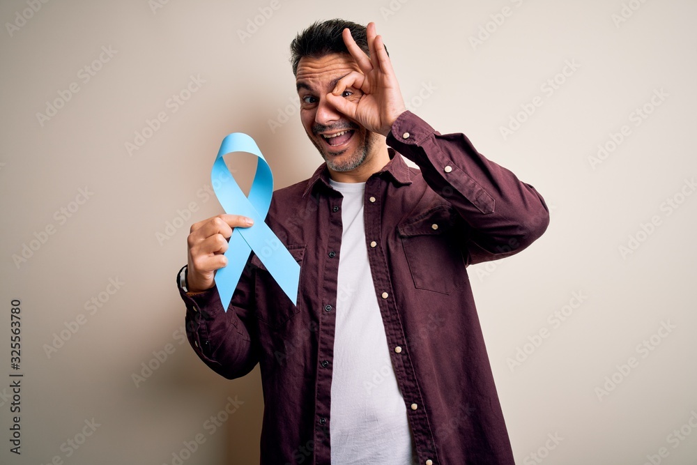 Young handsome man holding blue cancer ribbon over isolated white background with happy face smiling doing ok sign with hand on eye looking through fingers Young handsome man holding blue cancer ribbon over isolated white background with happy face smiling doing ok sign with hand on eye looking through fingers