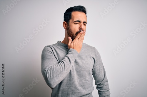 Young handsome man with beard wearing casual sweater standing over white background bored yawning tired covering mouth with hand. Restless and sleepiness.