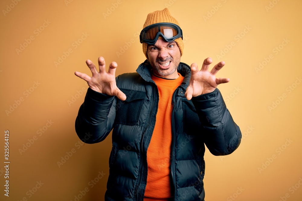 Young handsome skier man skiing wearing snow sportswear using ski goggles smiling funny doing claw gesture as cat, aggressive and sexy expression
