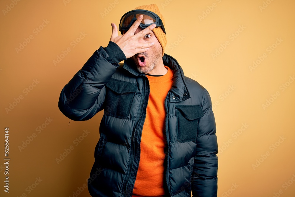 Young handsome skier man skiing wearing snow sportswear using ski goggles peeking in shock covering face and eyes with hand, looking through fingers with embarrassed expression. Young handsome skier man skiing wearing snow sportswear using ski goggles peeking in shock covering face and eyes with hand, looking through fingers with embarrassed expression.