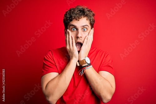 Young blond handsome man with curly hair wearing casual t-shirt over red background afraid and shocked, surprise and amazed expression with hands on face